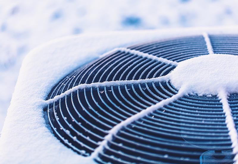 A close-up, top-down view of an outdoor HVAC or air conditioning unit unit covered in a layer of fresh white snow and frost, with snow clinging to the circular top fan grille.