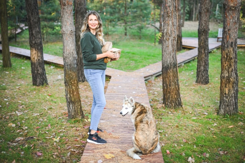 A woman standing on a wooden walkway in a wooded park with her husky sitting beside her.