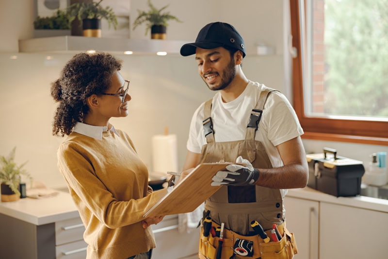 A home service technician reviewing paperwork on a clipboard with a homeowner in her kitchen.