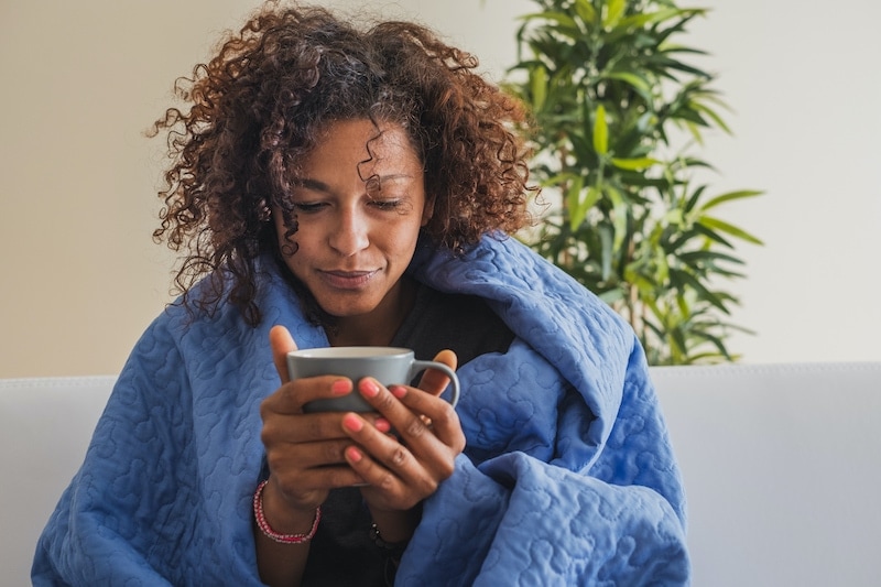 Woman is cold in her home so she is wrapped in a blue blanket and is drinking a mug of tea.